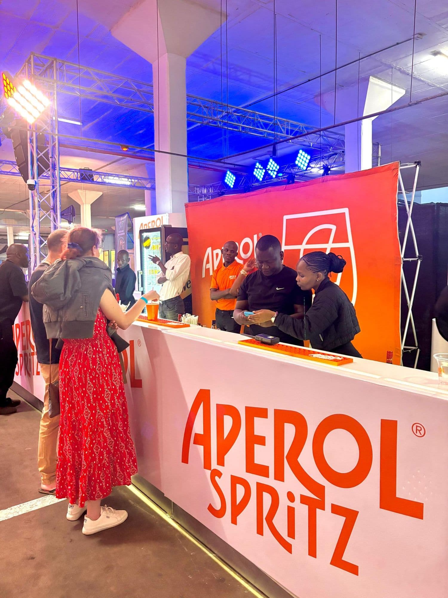An Aperol Spritz bar at an indoor event: a white counter carrying the Aperol Spritz wordmark, three crew members serving behind it against an orange backdrop with the Aperol glass roundel, and guests ordering at the near end