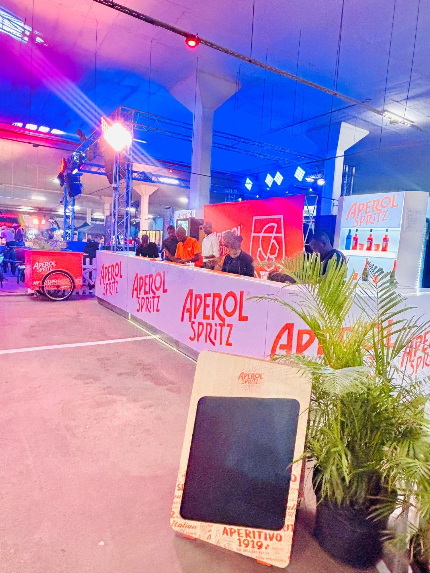 A long Aperol Spritz bar run under a lighting rig at an indoor event, the white counter carrying the wordmark four times over, with guests served along it, a branded trolley at the far end and a lit bottle display beyond