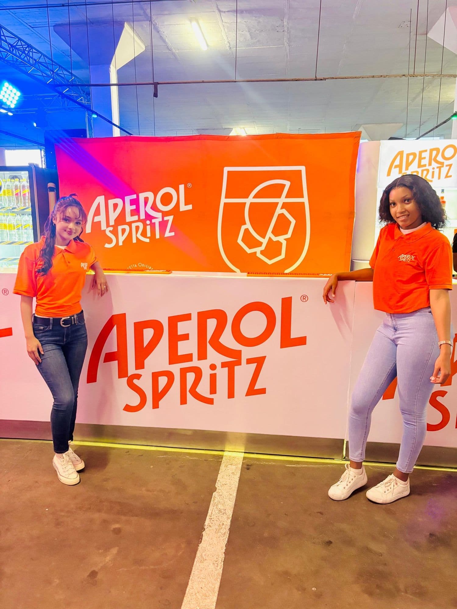 Two promoters in Aperol Spritz branded polo shirts standing either side of the branded bar counter, the wordmark on the counter front and the glass roundel on the backdrop behind them