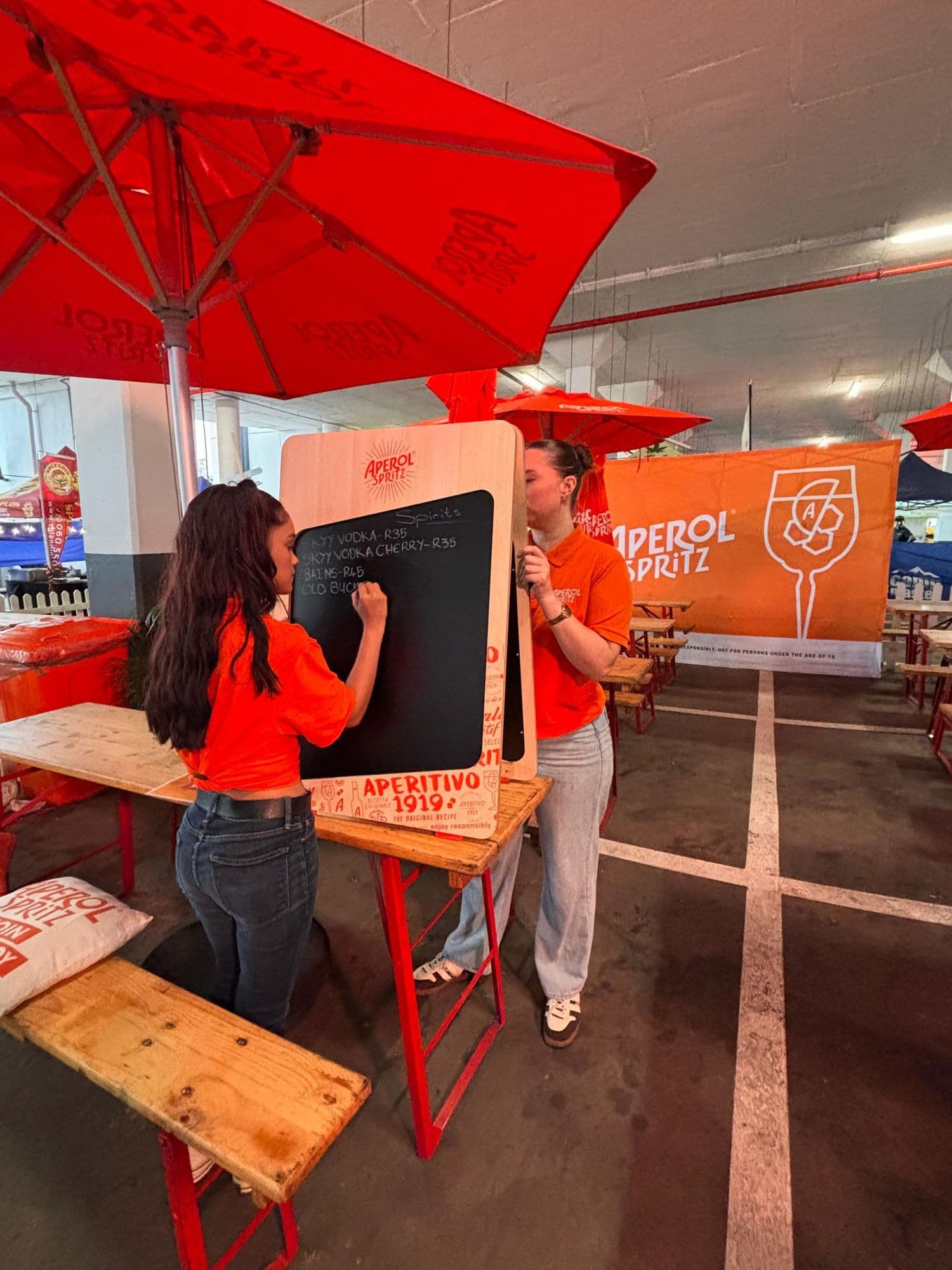 Two crew in Aperol Spritz branded kit setting up before doors: one chalking the spirits list onto a branded A-frame board while the other steadies it, under branded parasols with an Aperol Spritz banner behind them