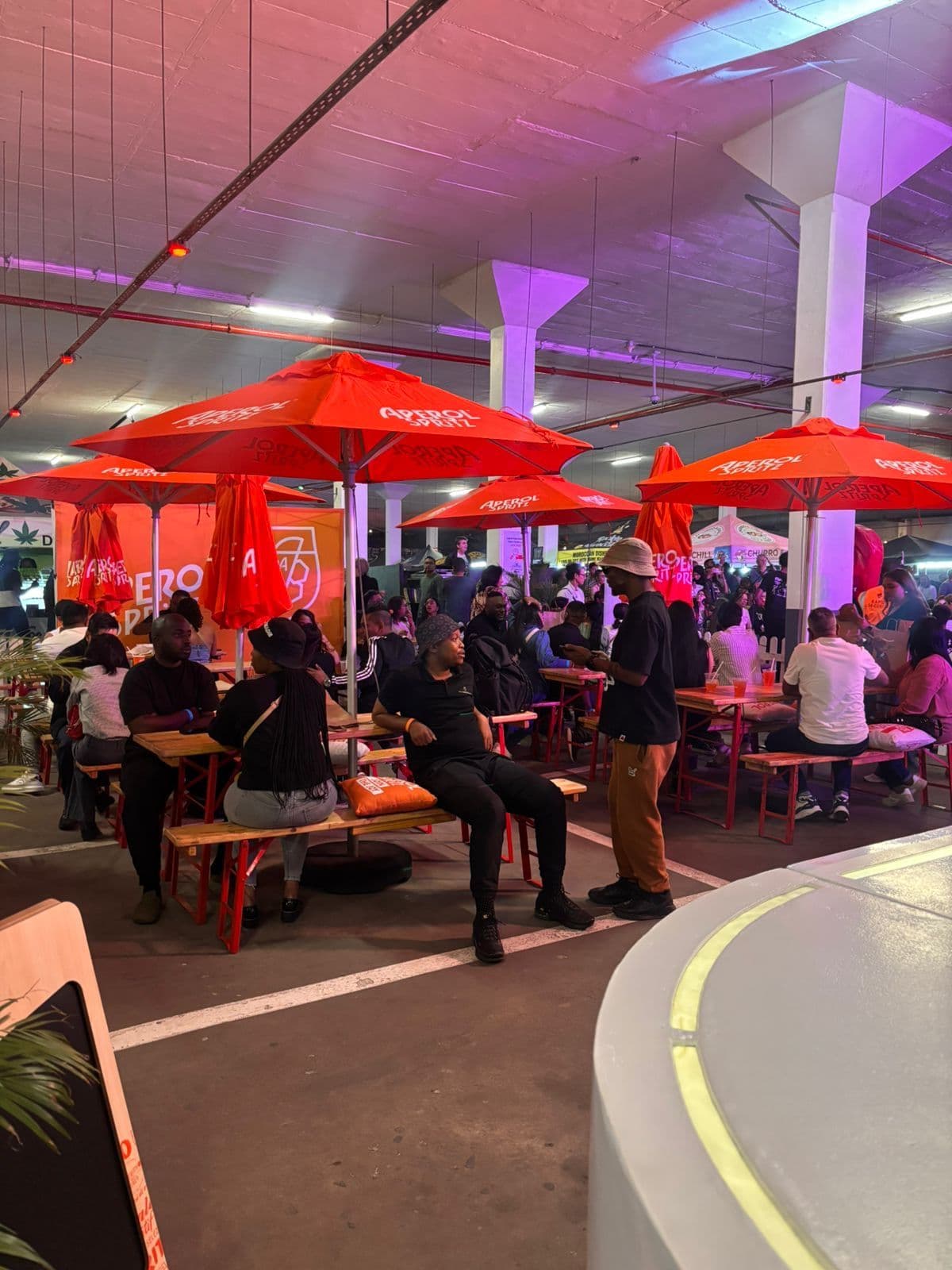 A busy Aperol Spritz terrace at an indoor event: a run of red branded parasols over long benches, with guests seated and drinking beneath them and a branded backdrop at the far end