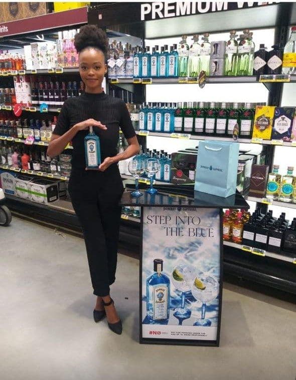 A promoter presenting the bottle between both hands under the premium white spirits sign, the tasting table set behind her