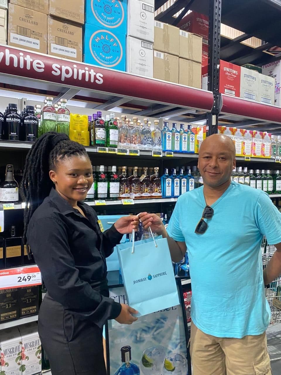 A promoter handing the turquoise Bombay Sapphire gift bag to a shopper, his partner alongside, gin shelves behind