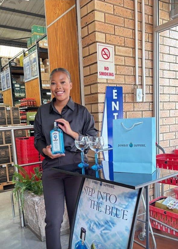 A promoter with a bottle of Bombay Sapphire at a black tasting table set with copa glasses and a gift bag, the banner beside a brick pillar