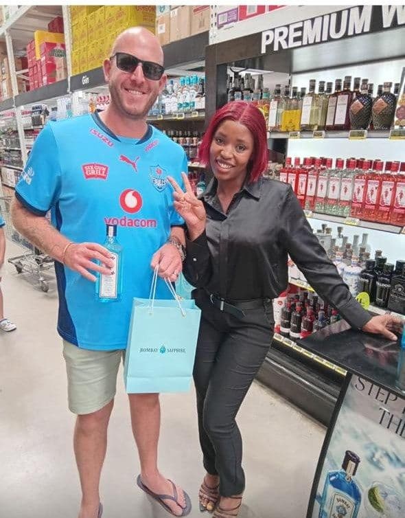 A shopper in a blue rugby shirt holding his gin and gift bag, the promoter beside him flashing a peace sign in the Makro aisle