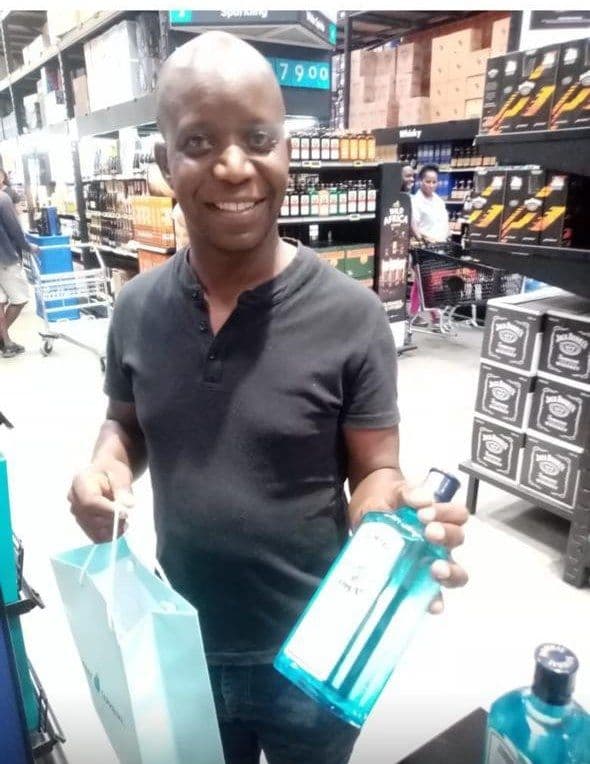 A grinning shopper with the bottle in one hand and the gift bag in the other, the whisky shelves behind him