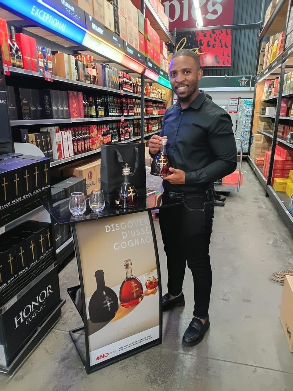 A promoter holding a bottle of D'Ussé beside the Discover D'Ussé banner stand in the brandy aisle, Honor Cognac cases stacked alongside