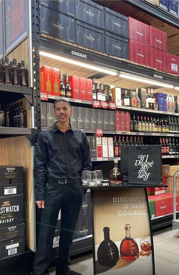 A promoter beside the tasting table under the store's cognac sign, the D'Ussé gift bag on the table and the Discover banner at his side