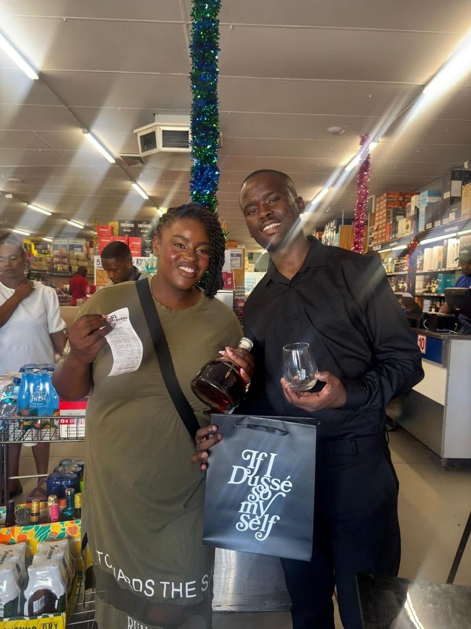 A shopper holding her till slip and a tasting glass, the promoter beside her with the gift bag, tinsel overhead