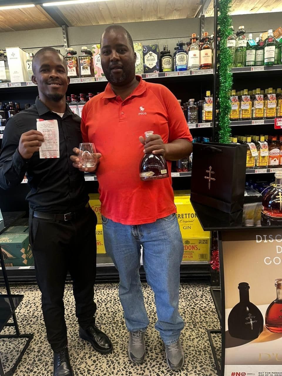 A promoter and a shopper in a red shirt at the shelf, the shopper holding his bottle of D'Ussé and a tasting glass