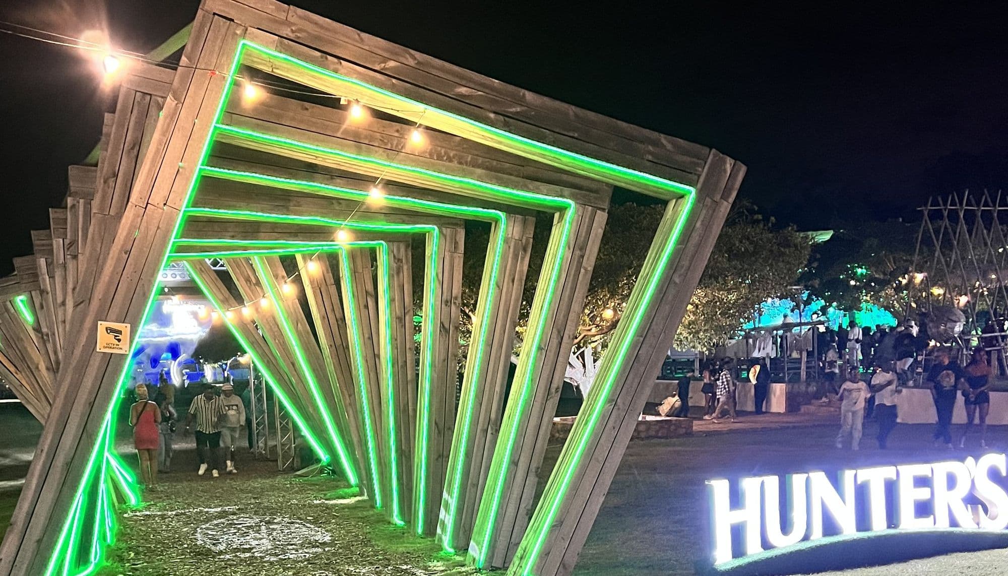 The same entrance tunnel built on site at night: the tilted timber frames traced in green neon with festoon lights strung between them, guests walking through, and an illuminated HUNTER’S sign standing on the grass beside it
