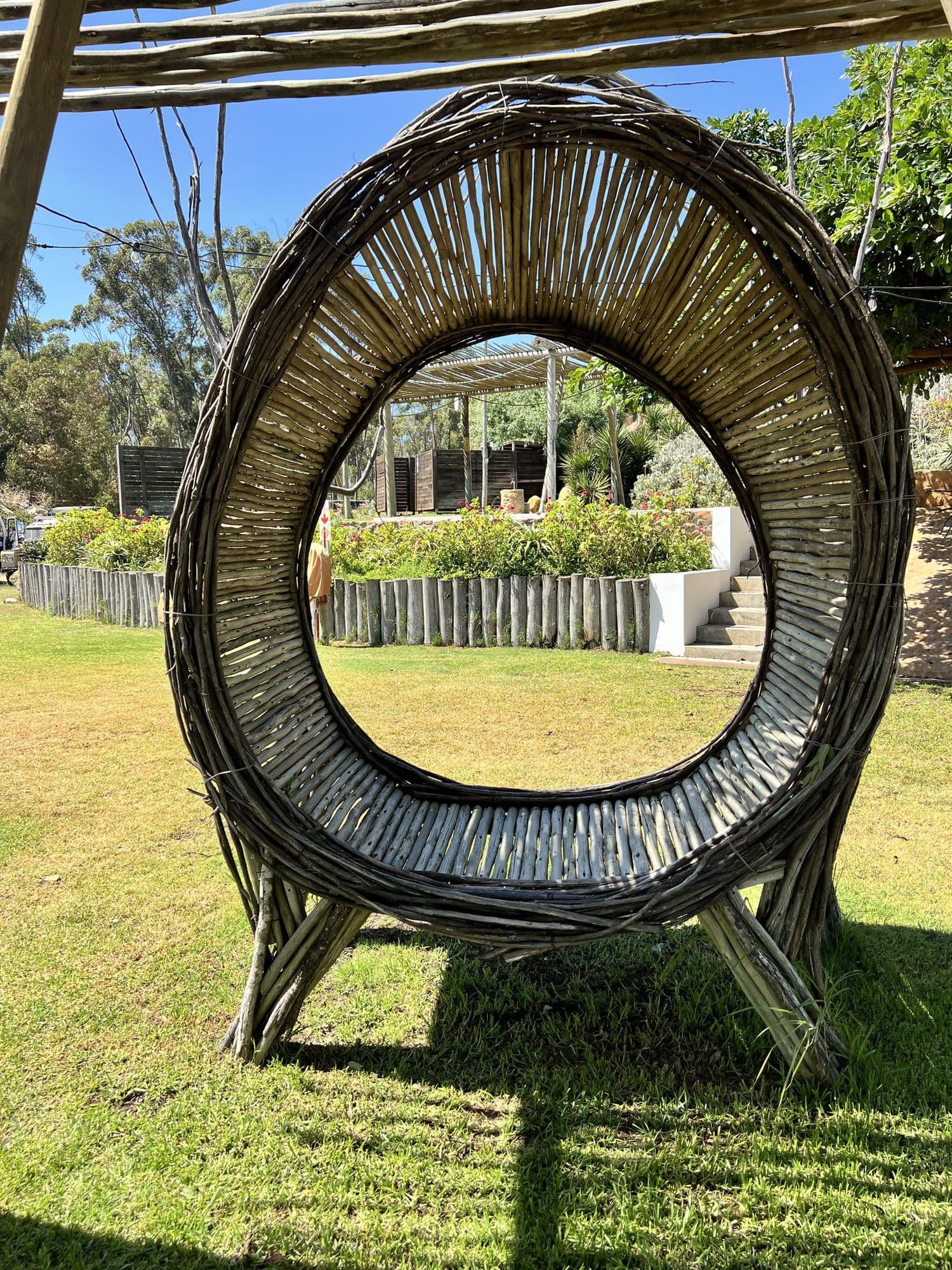 A circular pod woven from timber branches standing bare on a lawn in daylight, its open centre framing a garden bed and steps beyond, before any dressing has been applied