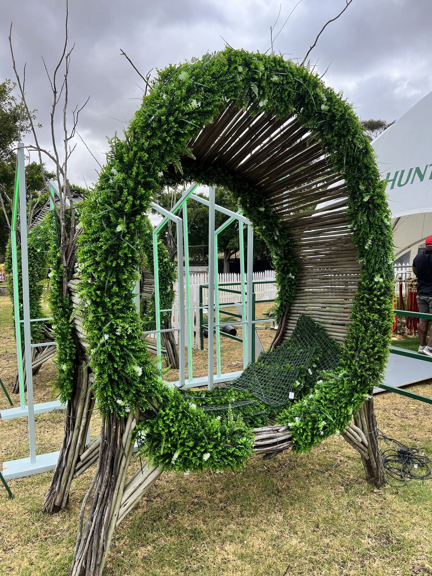 The same woven ring pod on site the next day, dressed with dense green foliage around its rim, standing beside neon-lit frames with a Hunter’s marquee behind it