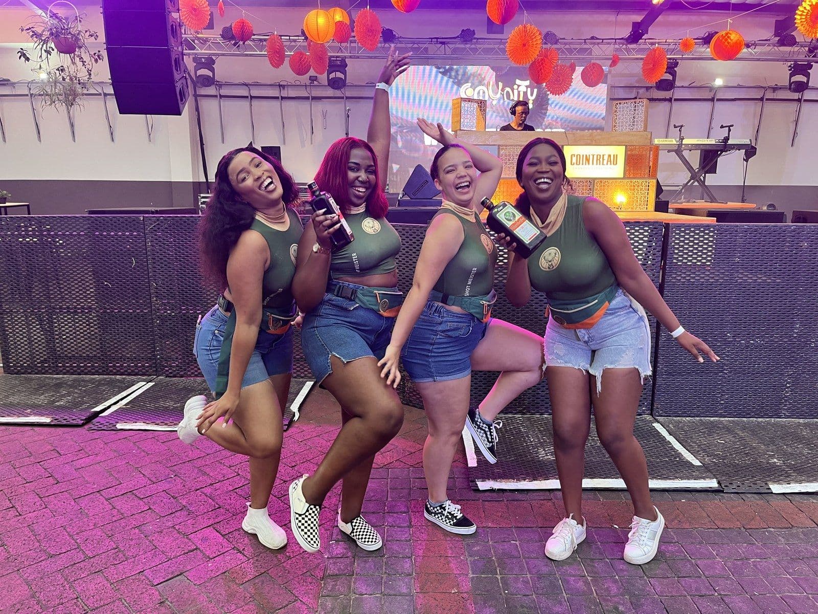 Four Jägermeister ambassadors dancing with bottles in hand at an indoor event, orange paper lanterns and a DJ booth behind them