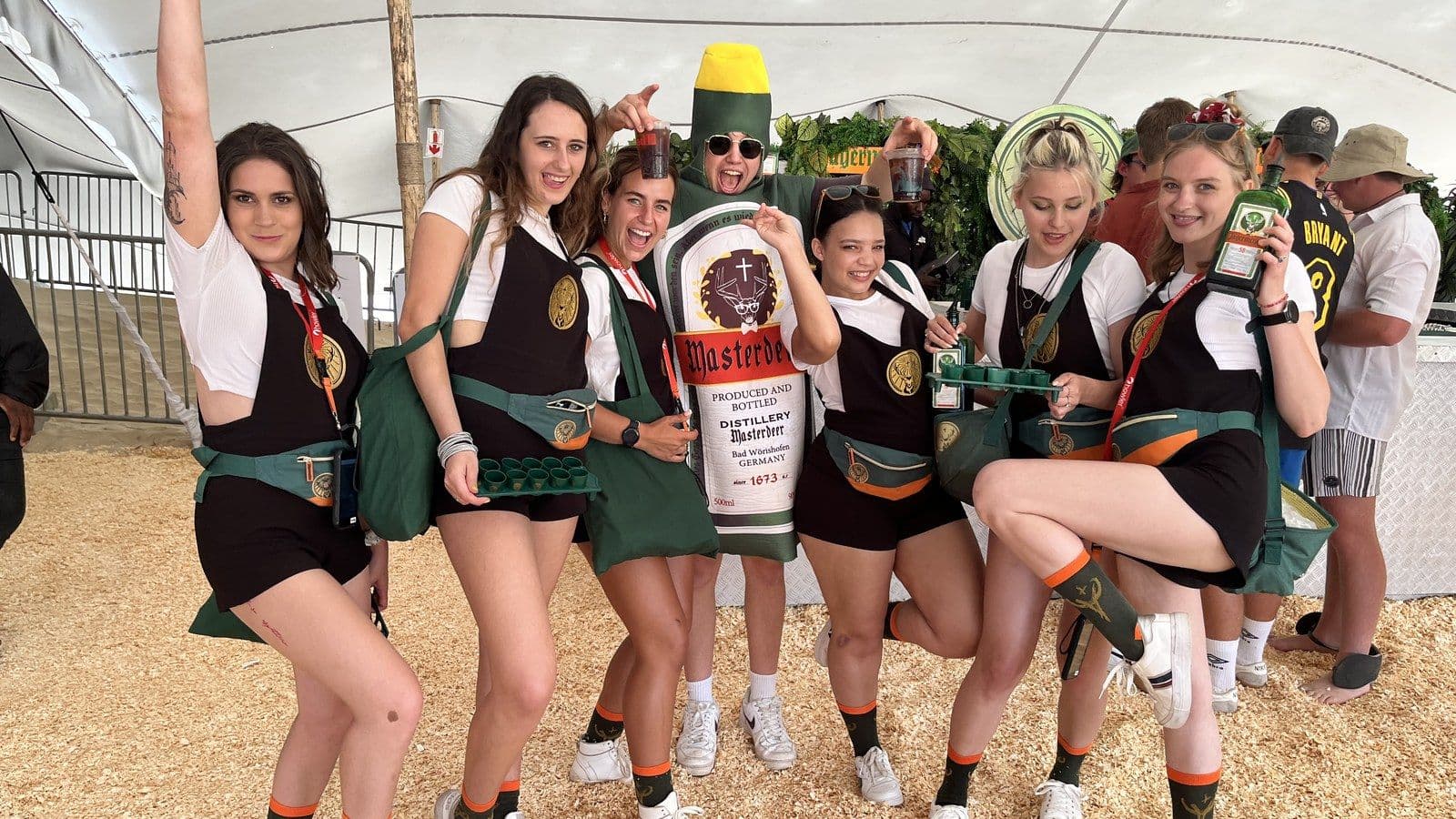 Six Jägermeister brand ambassadors in kit with shot trays and bottles, laughing around a person in a Jägermeister bottle costume, under a festival stretch tent