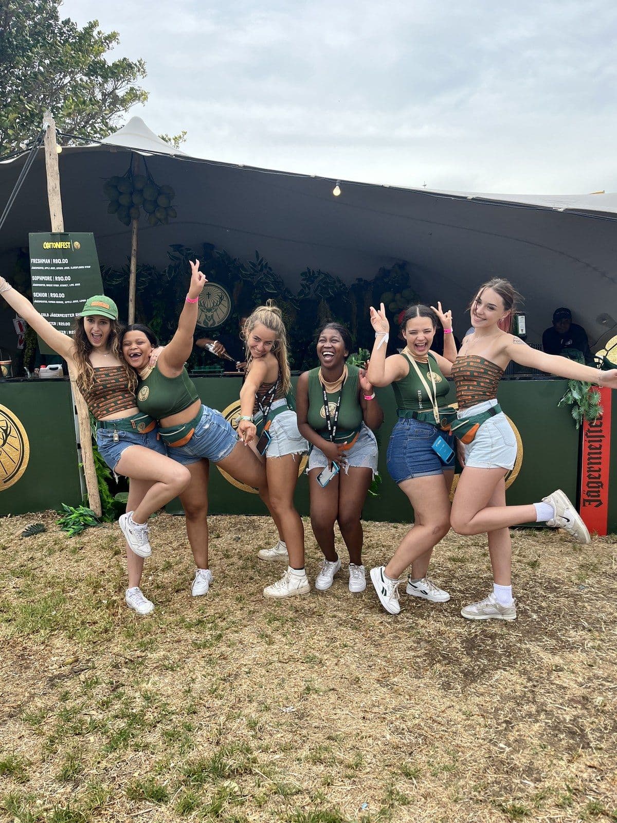 Six Jägermeister crew mid-pose on a festival field in front of the branded bar, arms up, one leg kicked out