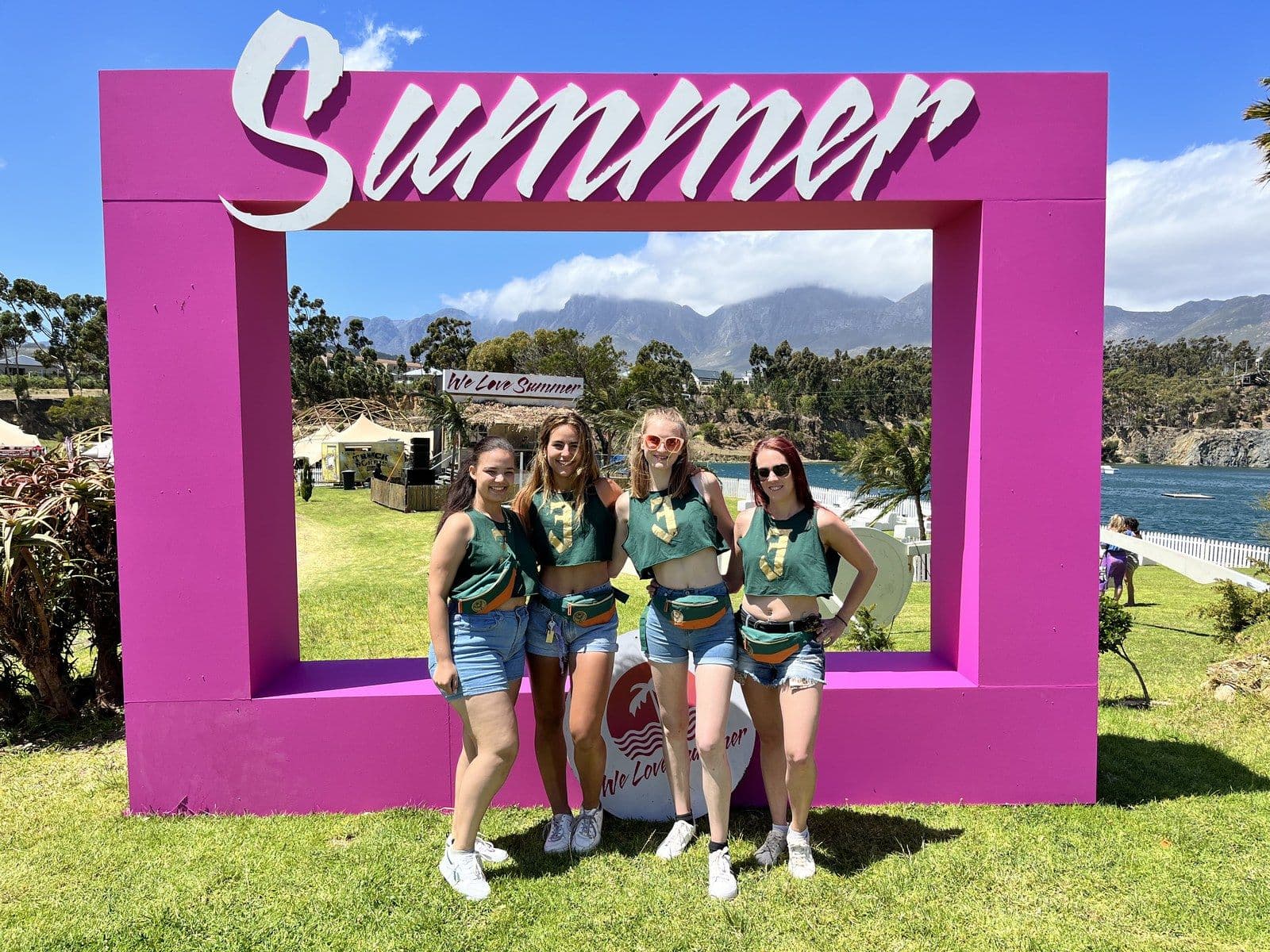 Four Jägermeister ambassadors posing inside a giant pink Summer photo frame on a lawn, mountains and the festival grounds behind them
