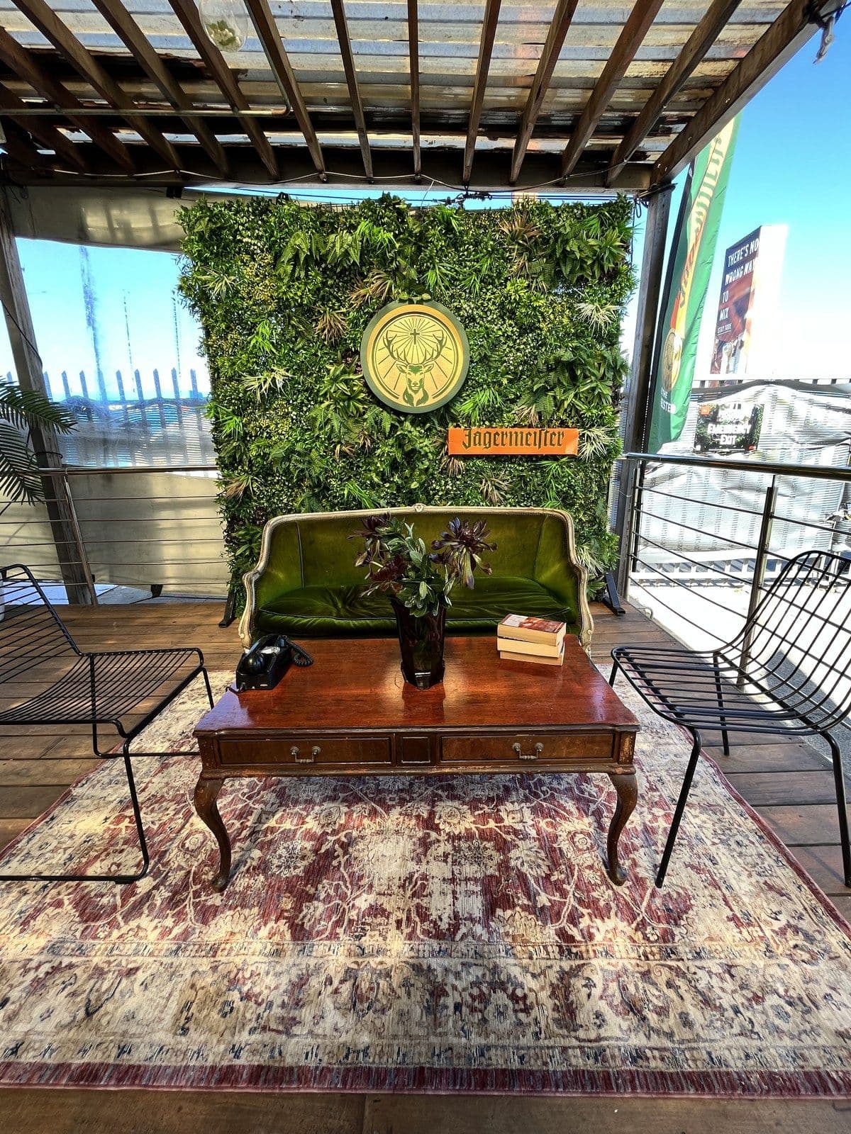 A styled Jägermeister terrace lounge — living green wall with the stag roundel and Jägermeister sign, a green velvet sofa, and a vintage coffee table with a rotary phone and books