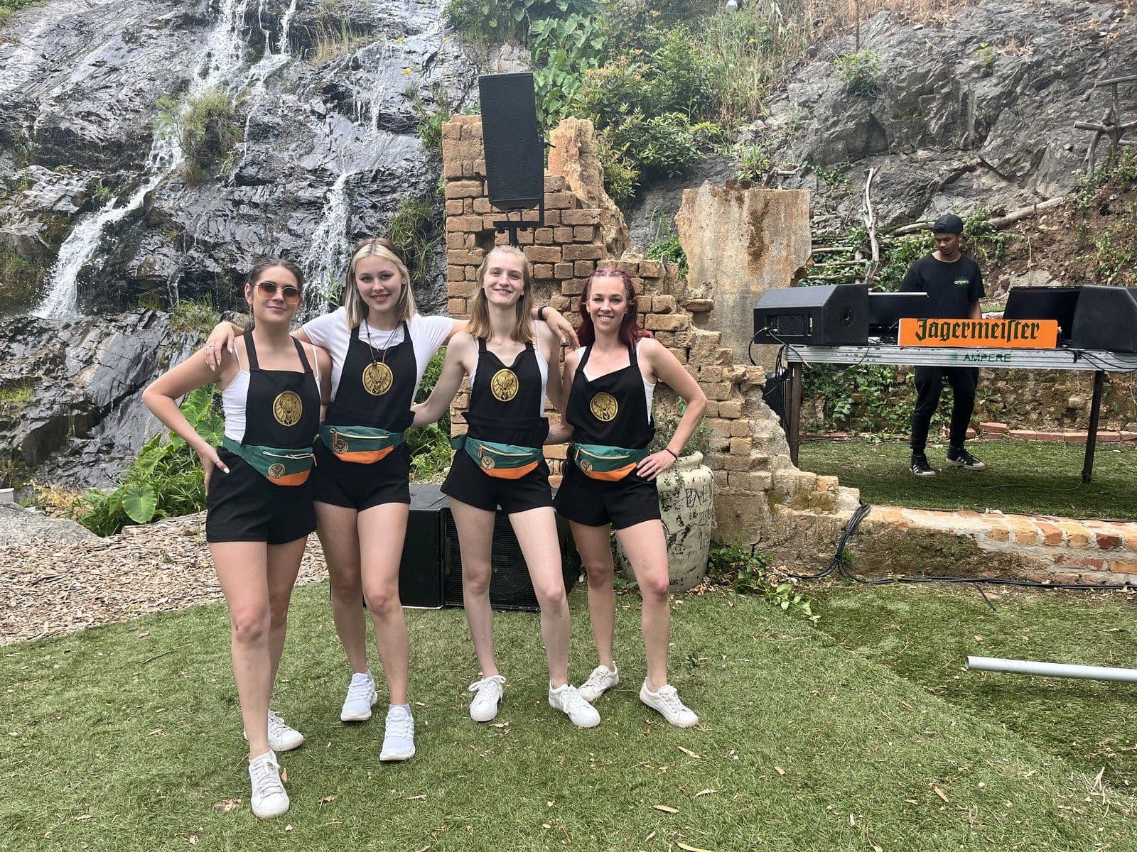 Four Jägermeister ambassadors in kit arm in arm on a lawn in front of a waterfall, a DJ playing beside them under an orange Jägermeister sign