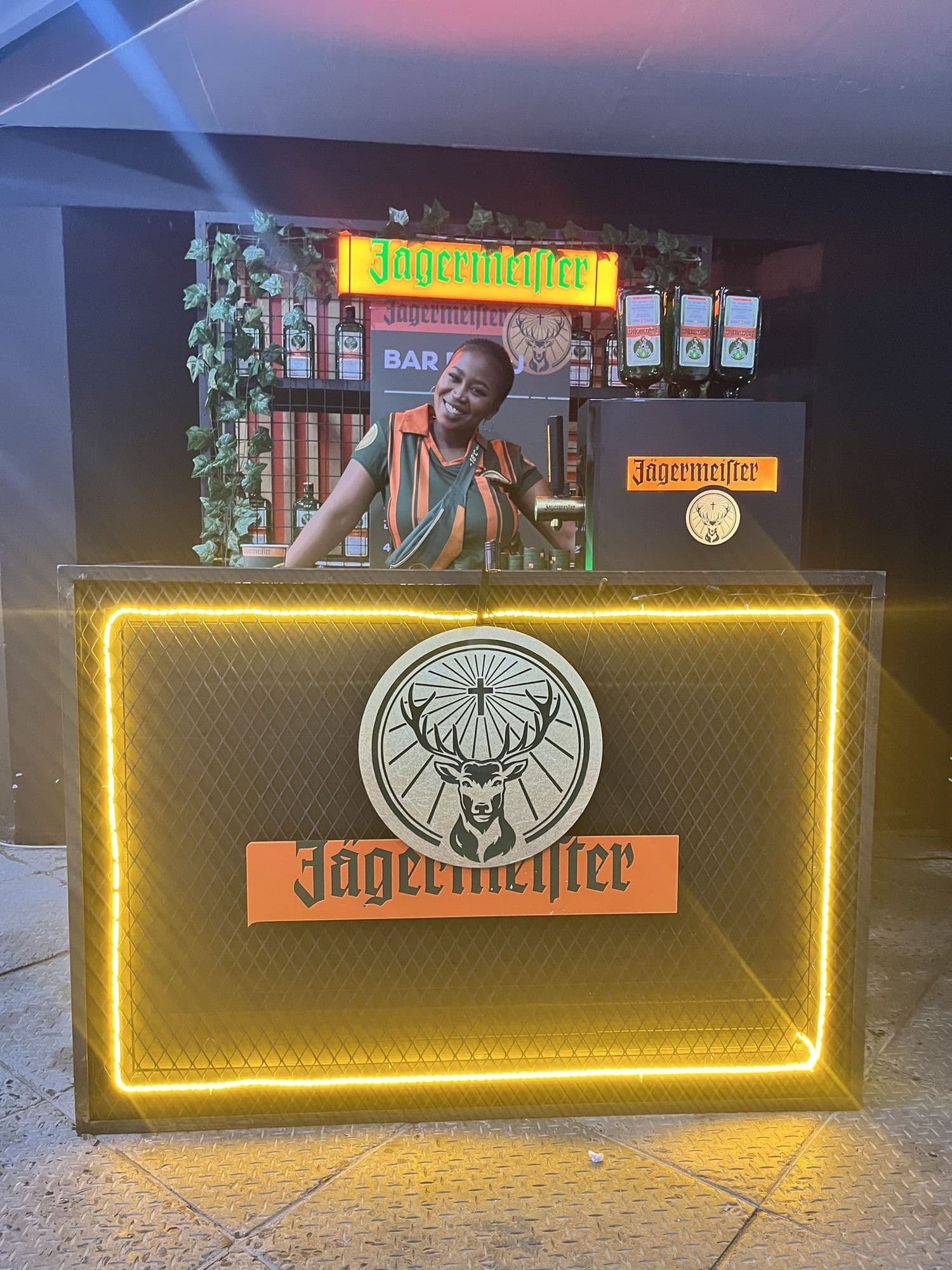 An illuminated Jägermeister branded bar unit with the stag roundel on the front panel, bottles on optics and a bartender in branded kit behind it