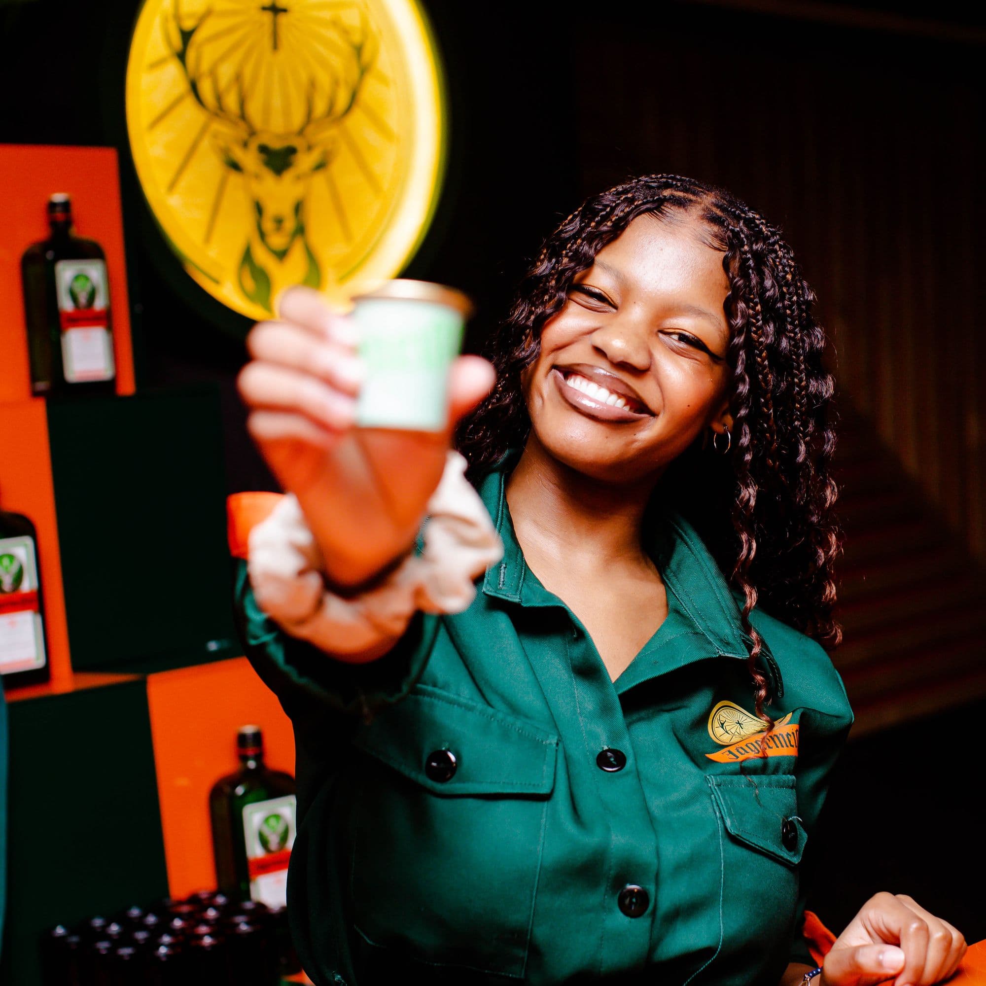 A Jägermeister ambassador in green kit holding a shot out to the camera, the stag roundel lit behind her