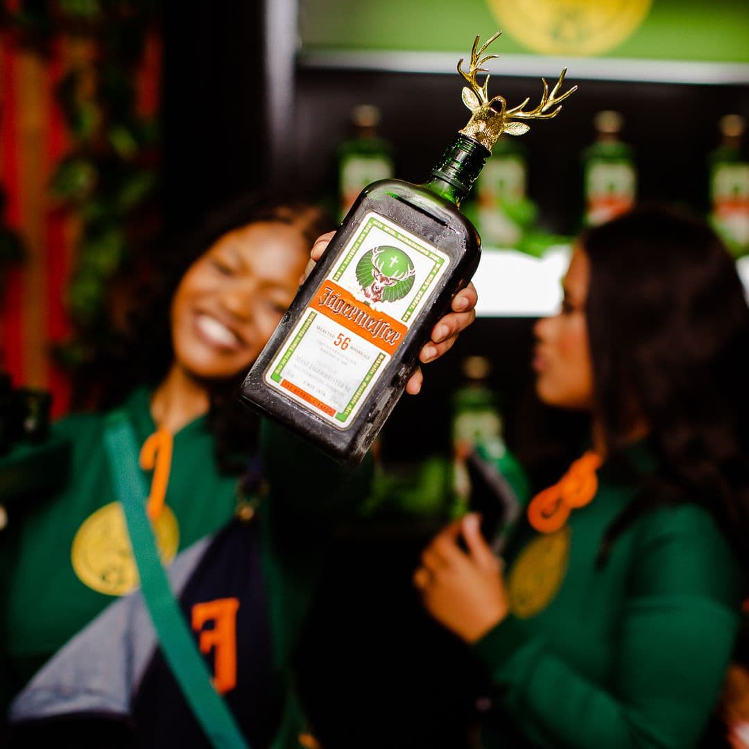 A frosted Jägermeister bottle with a gold stag topper held up at an on-trade activation, brand ambassadors behind it