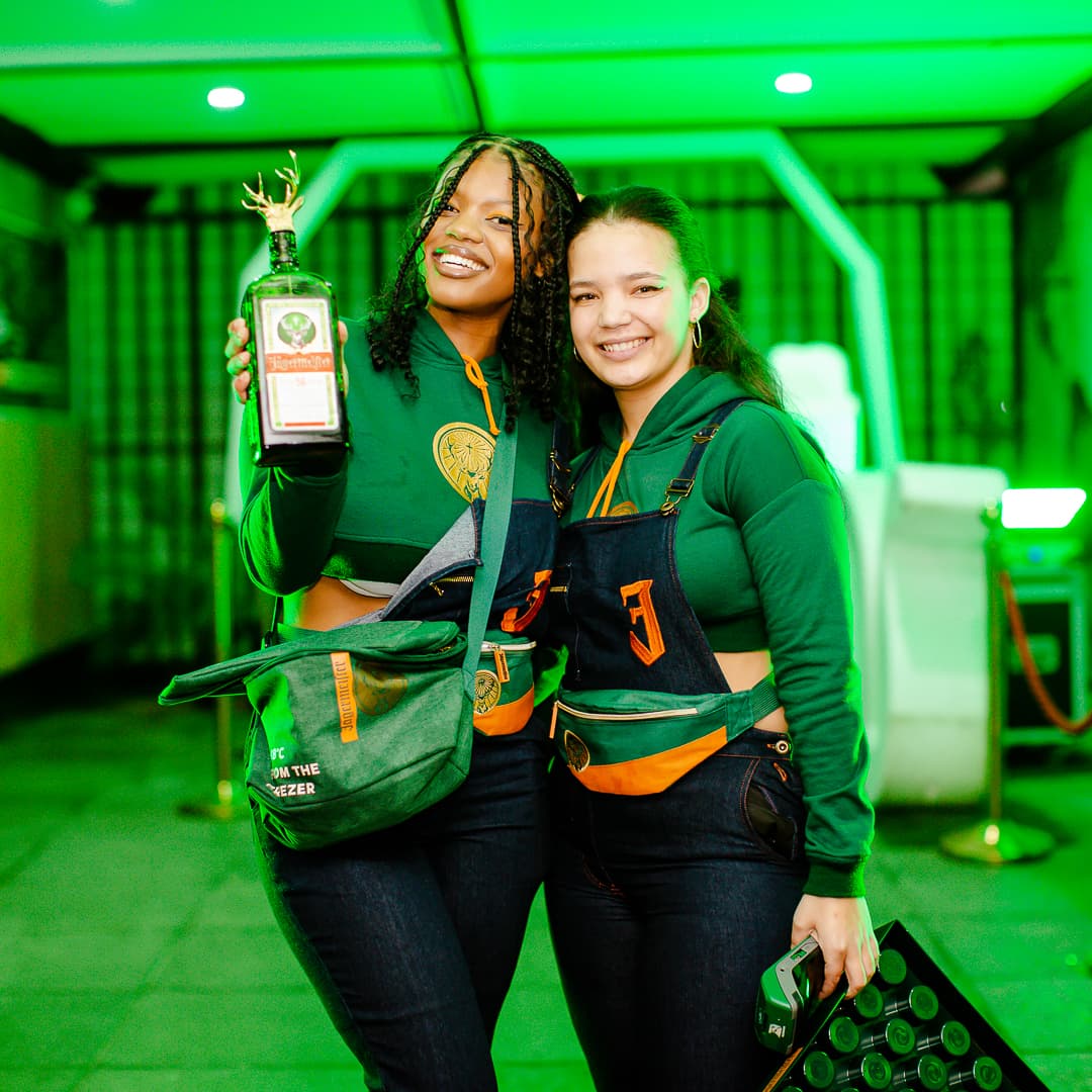 Two Jägermeister ambassadors in full kit, one presenting a frosted bottle, the other carrying a tray of shots
