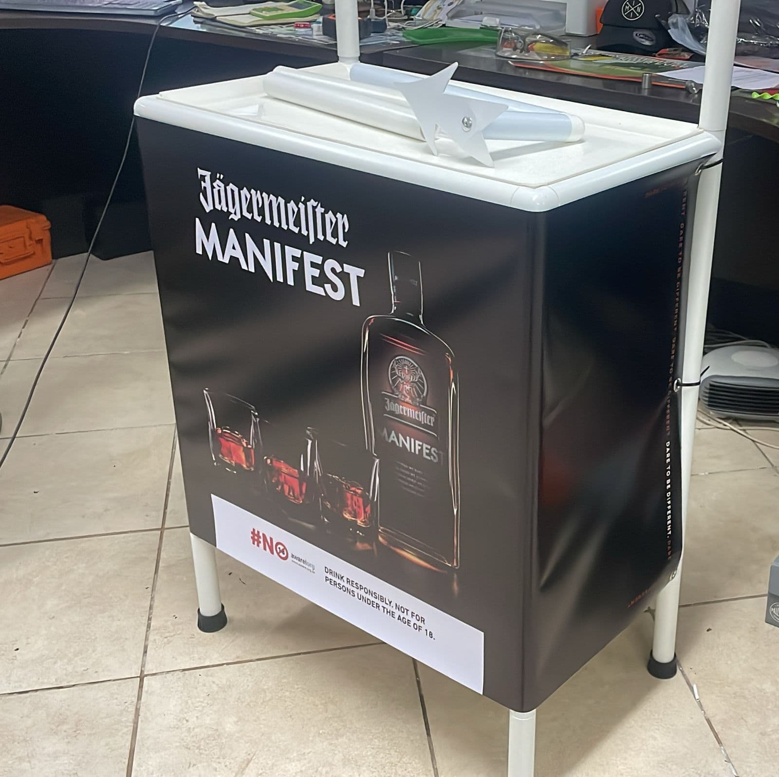 A branded Jägermeister Manifest ice bin on a tiled floor, its printed panel showing the bottle and serves alongside the responsible-drinking strip