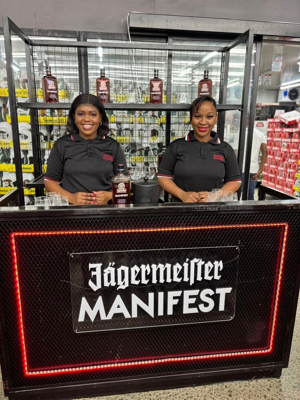 Two promoters in branded Jägermeister Manifest polo shirts standing behind an illuminated Manifest bar unit inside a liquor store, with Manifest bottles and tasting glasses on the shelf behind them
