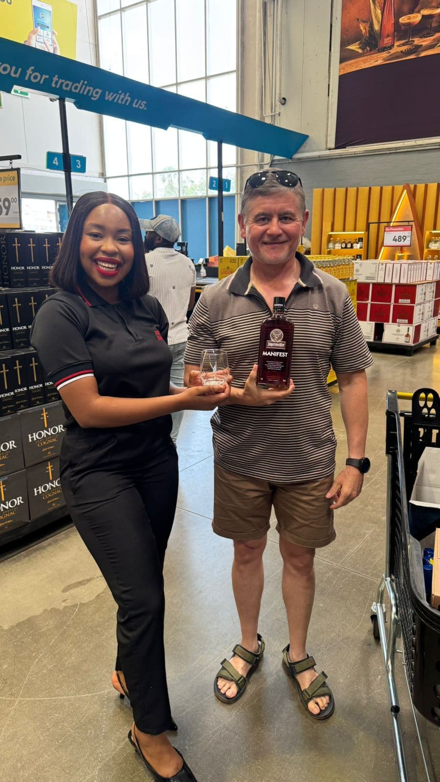 A promoter in Jägermeister Manifest kit handing a tasting glass to a shopper holding a Manifest bottle, on the floor of a warehouse liquor store