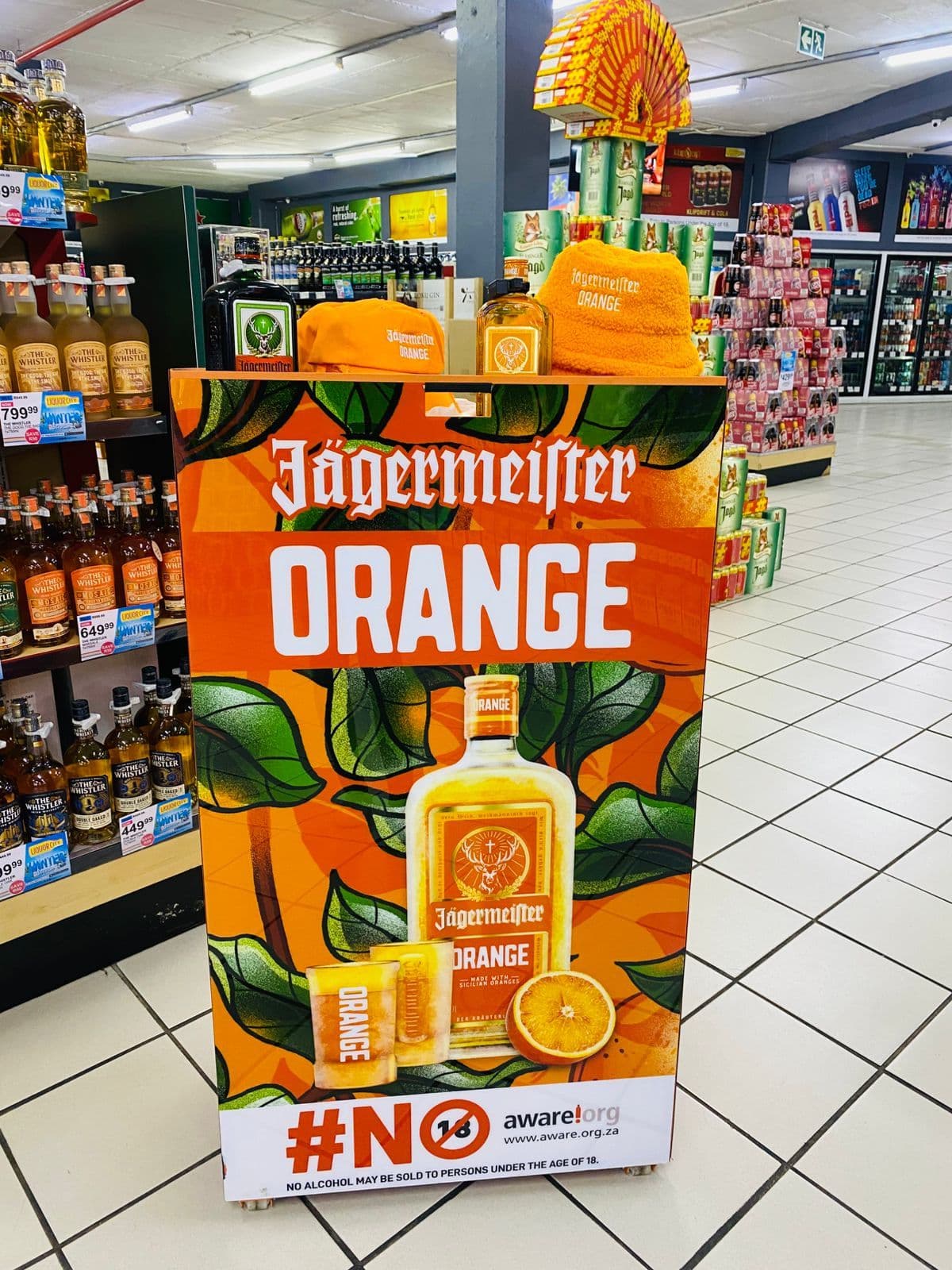A free-standing Jägermeister Orange display unit on a liquor store floor, printed with the bottle and orange-leaf artwork, with branded caps and a bucket hat merchandised on the shelf above it