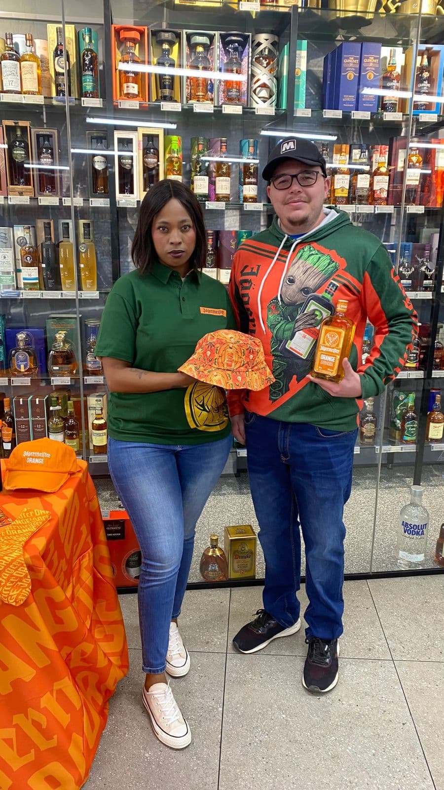 A promoter in Jägermeister branded kit handing a branded orange bucket hat to a shopper holding a bottle of Jägermeister Orange, in front of a lit spirits cabinet in a store