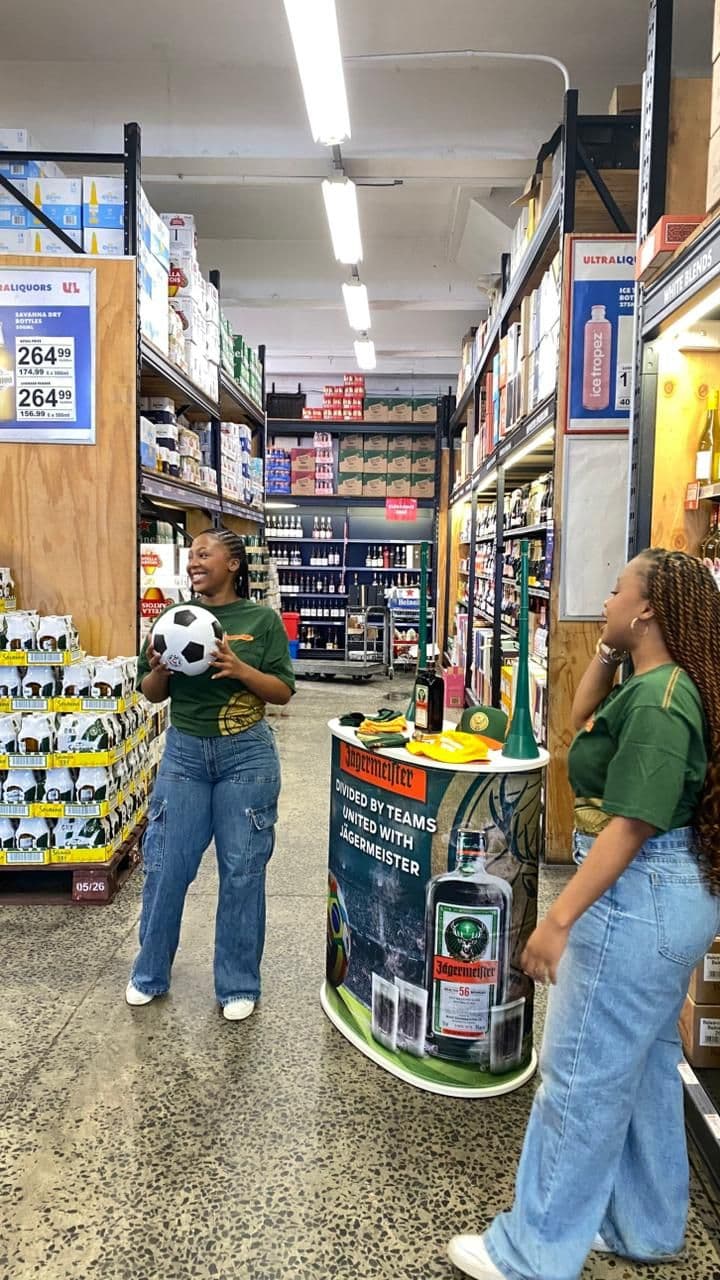 Two crew in branded green kit in a liquor store aisle beside the Jägermeister World Cup table, one holding a football, the table carrying the line “Divided by teams, united with Jägermeister”