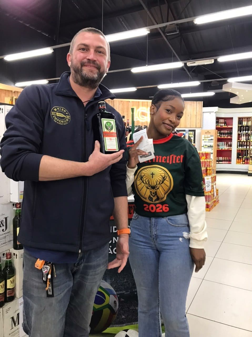 A bearded shopper holding his bottle beside a promoter in the 2026 campaign tee