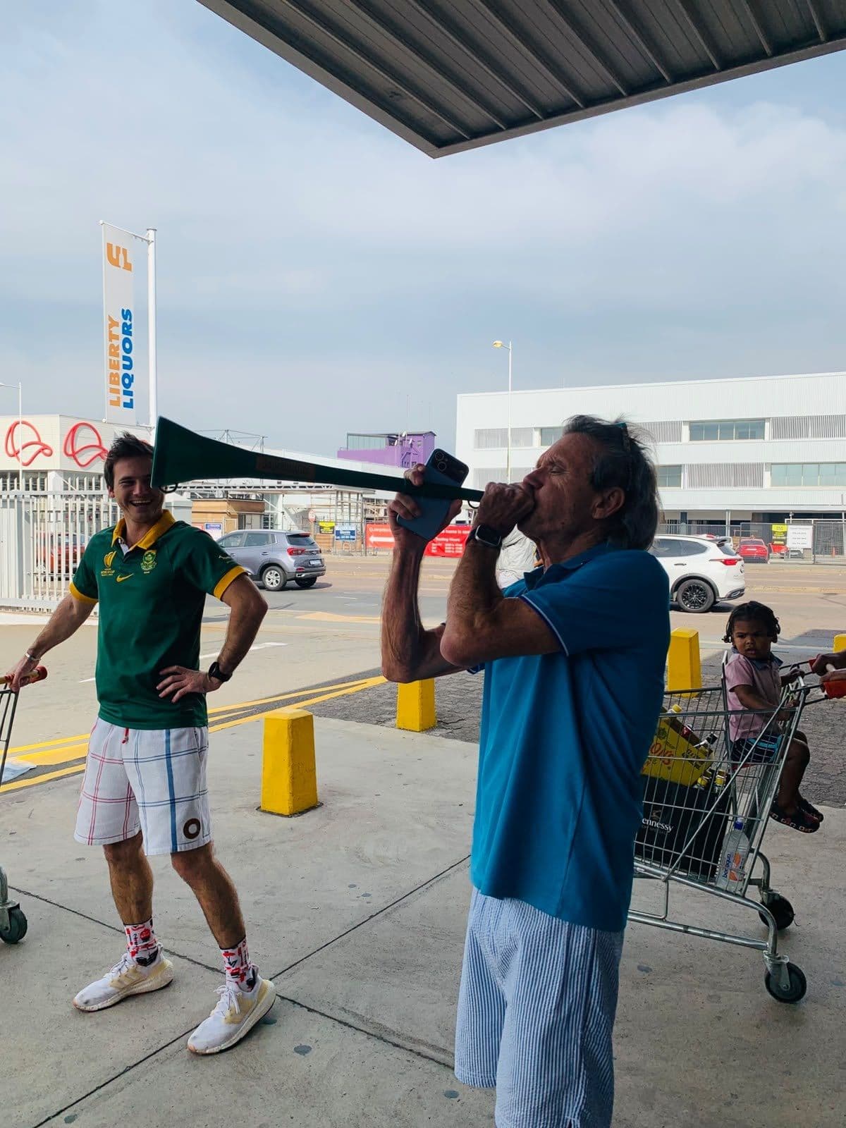A shopper testing the green vuvuzela on the store forecourt, the promoter with a flag beside him