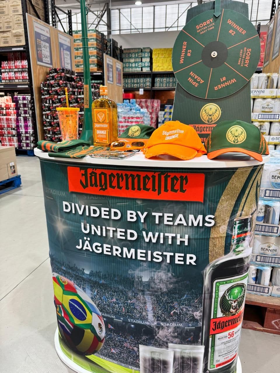 A Jägermeister World Cup sampling table in a warehouse liquor store, printed “Divided by teams, united with Jägermeister”, with a spin-to-win prize wheel above it and branded caps, sunglasses and a scarf laid out on the counter