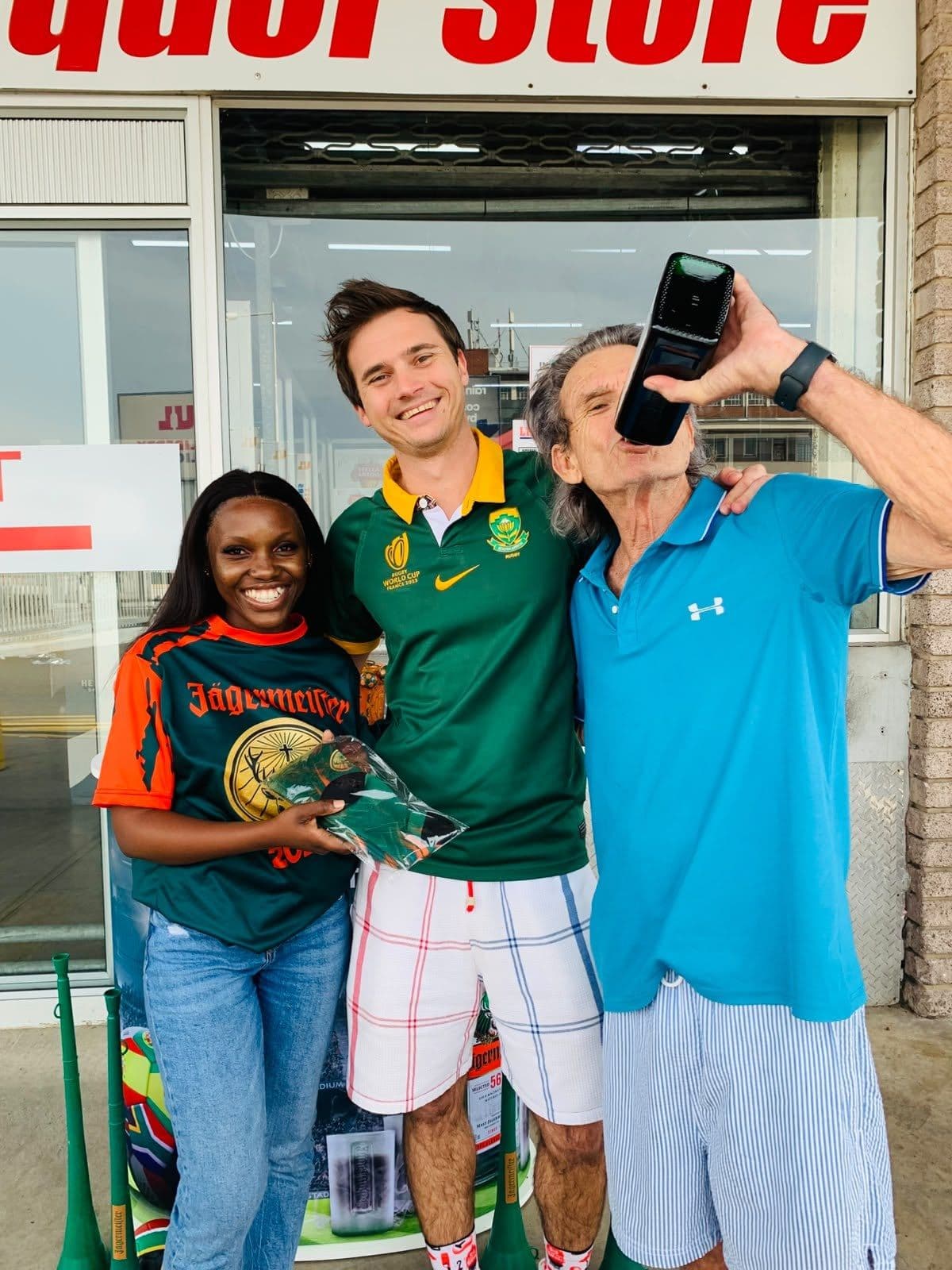 Three men posing outside the liquor store, one in a Springbok jersey, one raising a phone