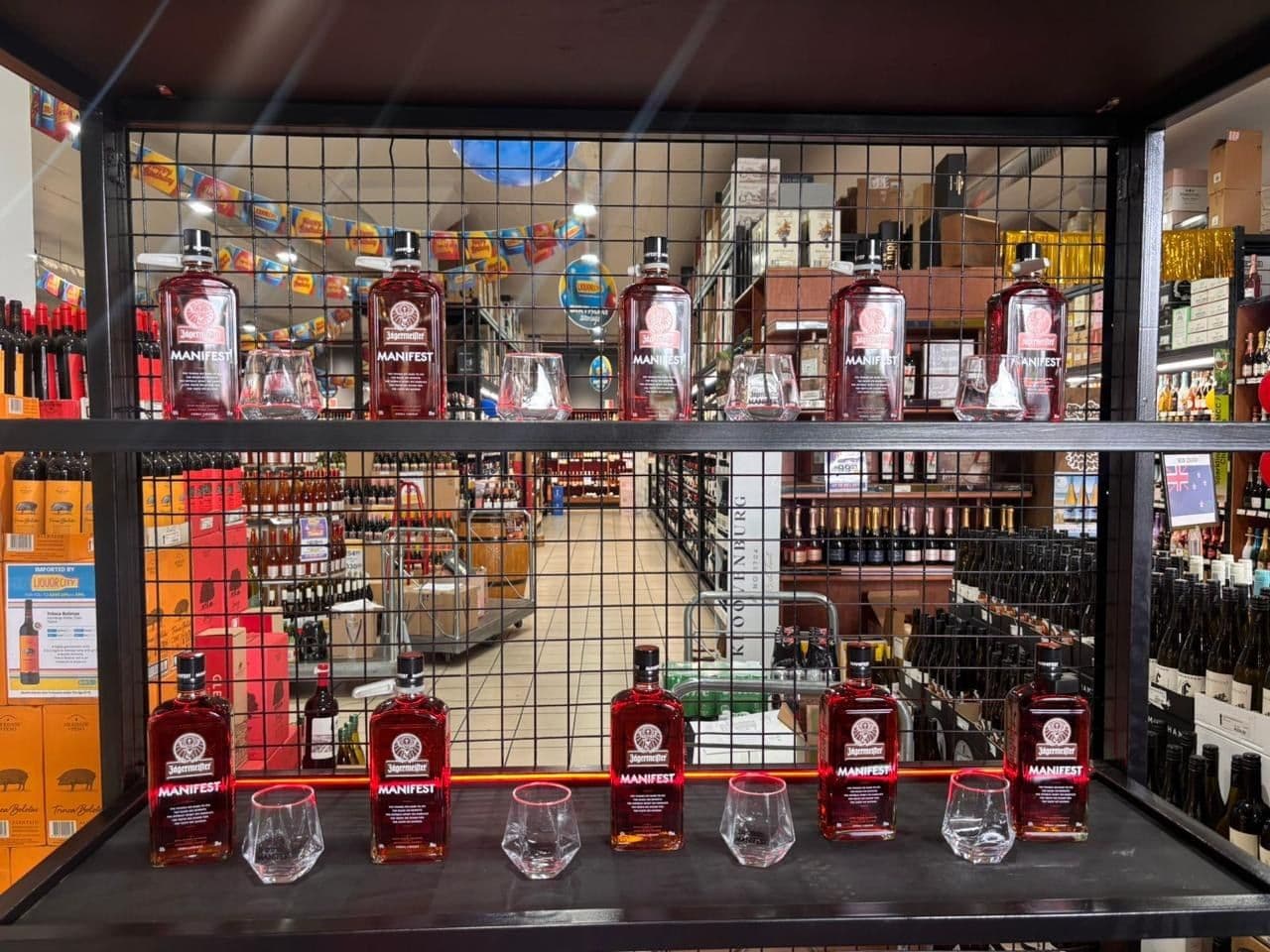 The back wall of the Jägermeister Manifest in-store bar: a black wire cage stocked with Manifest bottles and rows of tasting glasses