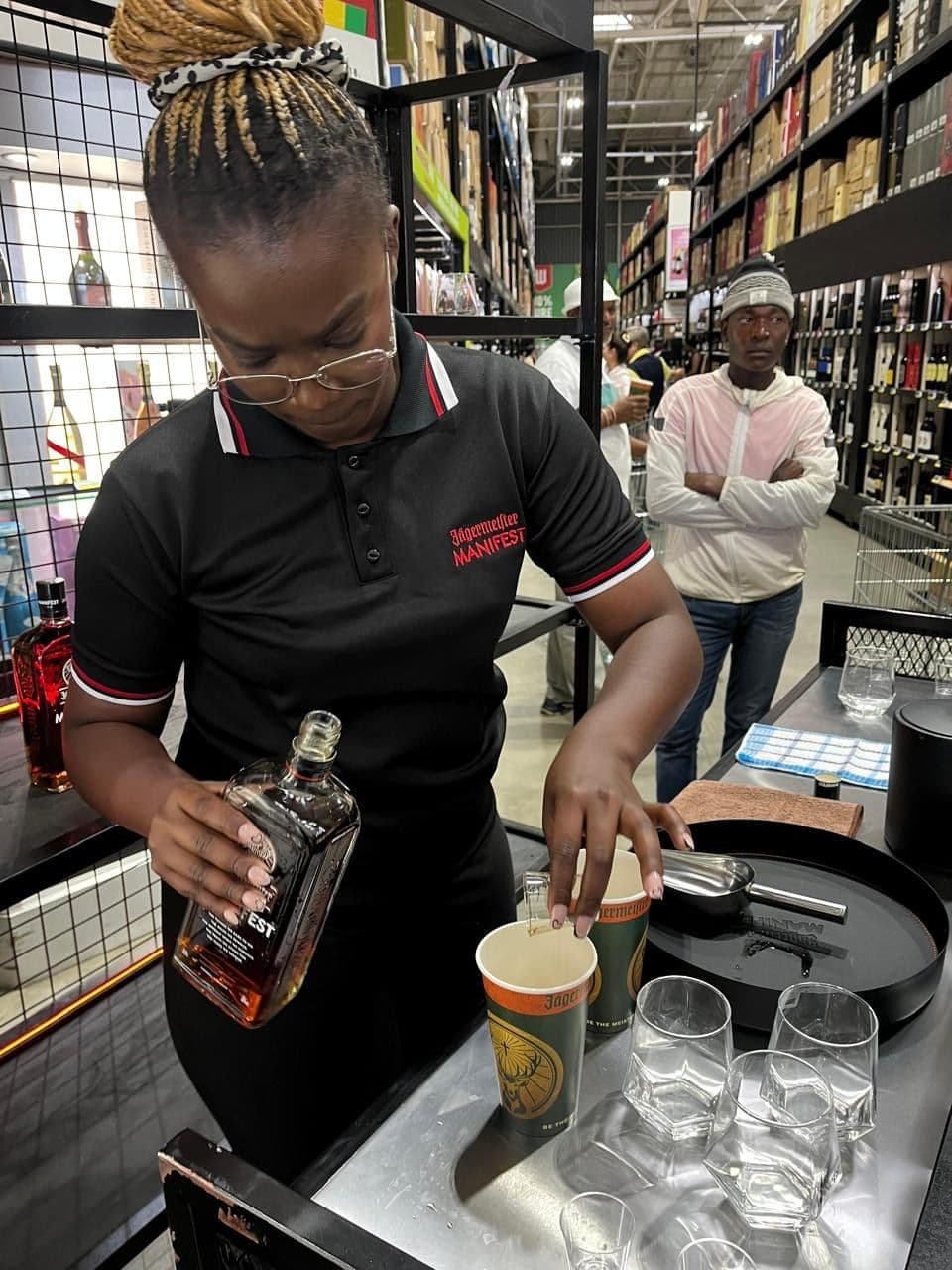 A promoter pouring Manifest into a serve over a tray of tasting glasses at the end of the bar
