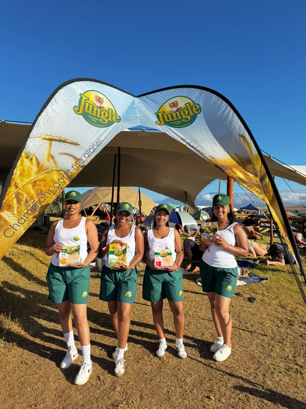A branded Jungle arch standing on grass at an outdoor event, printed with two Jungle tiger roundels and the line “Champions don’t rise alone”, with four promoters in branded caps, vests and shorts standing under it holding packs of Jungle oats and muesli