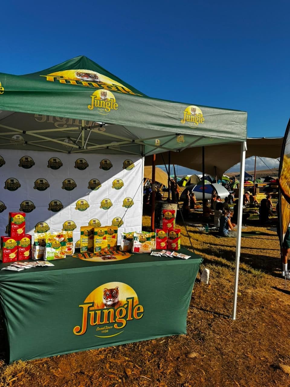 A Jungle branded gazebo at an outdoor event, its roof carrying the tiger roundel, a printed step-and-repeat backdrop behind and a skirted table laid out with oats, muesli and sachets