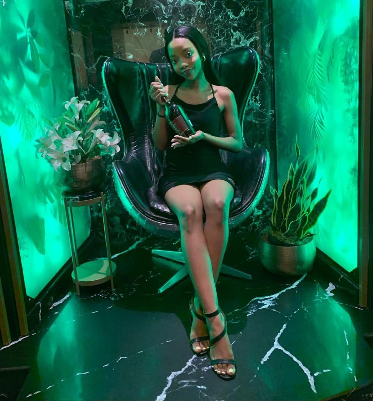 A model seated in a black egg chair with the bottle, against a green-lit marble wall dressed with plants