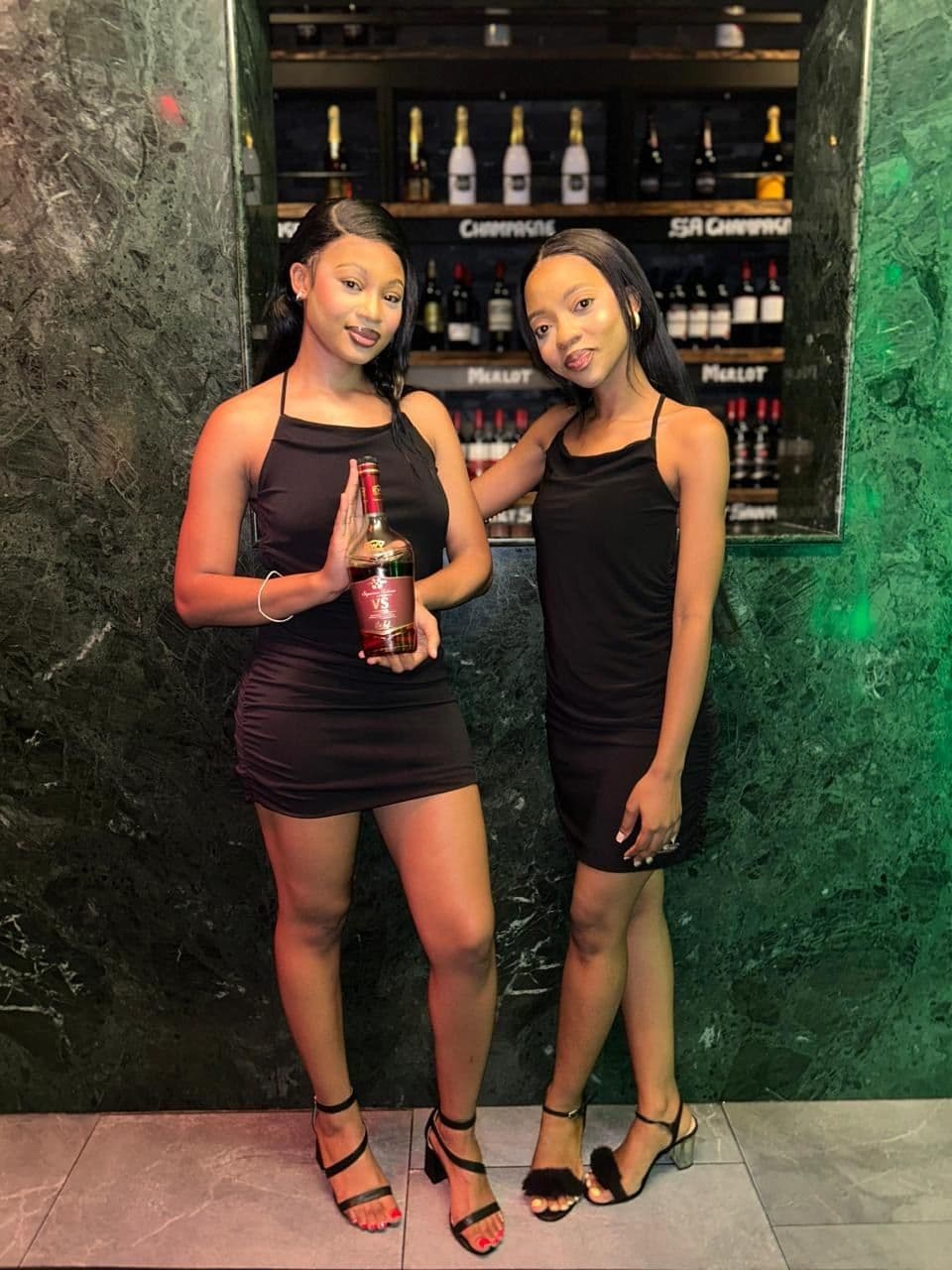 Two hostesses in black dresses, each holding a bottle of KWV VS, in front of the venue's wine wall