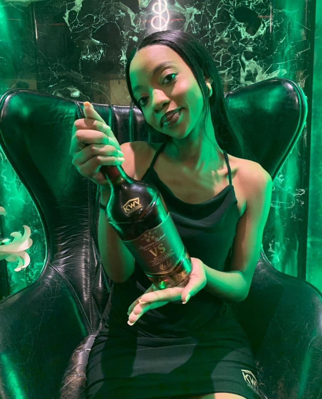 A model in the egg chair holding the bottle upright between both hands, label to camera
