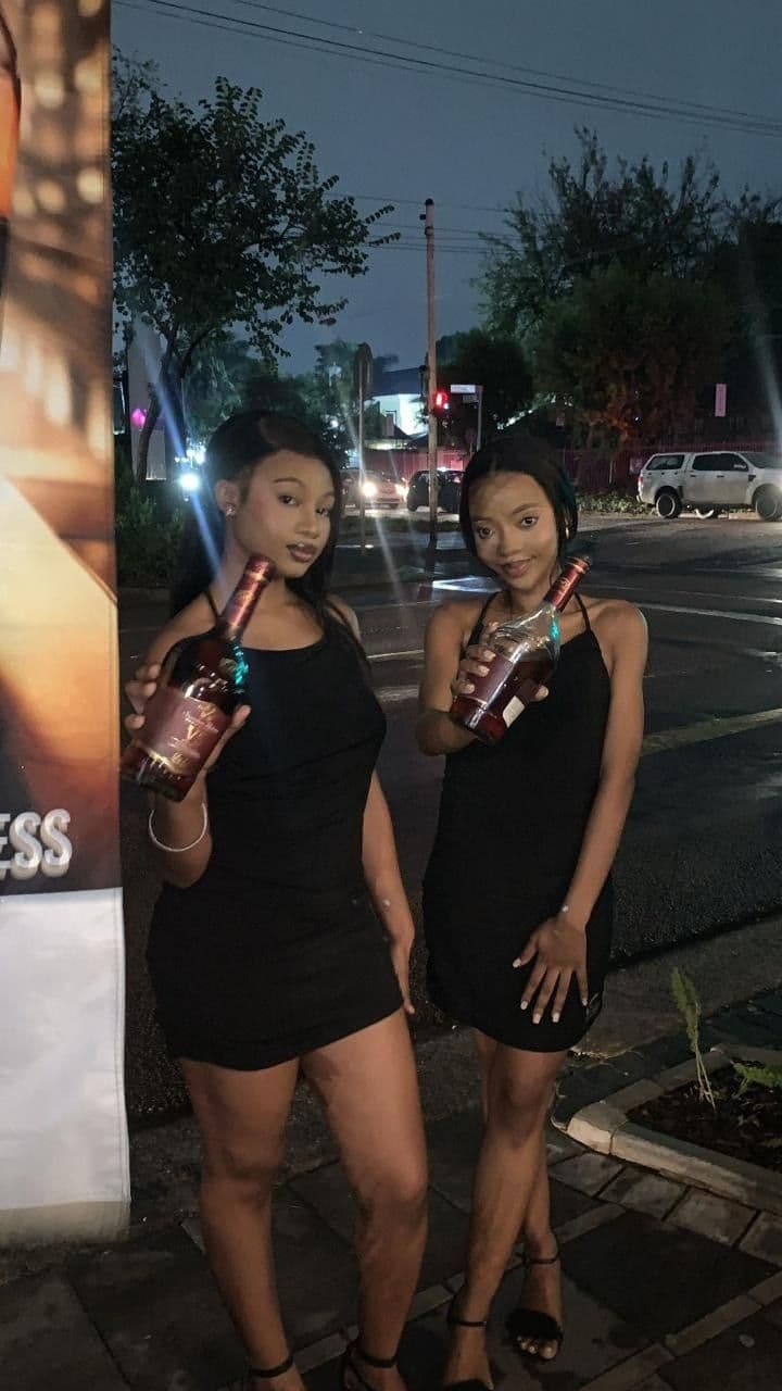 Two promoters with bottles out front of the venue at dusk, the street lights coming on behind them