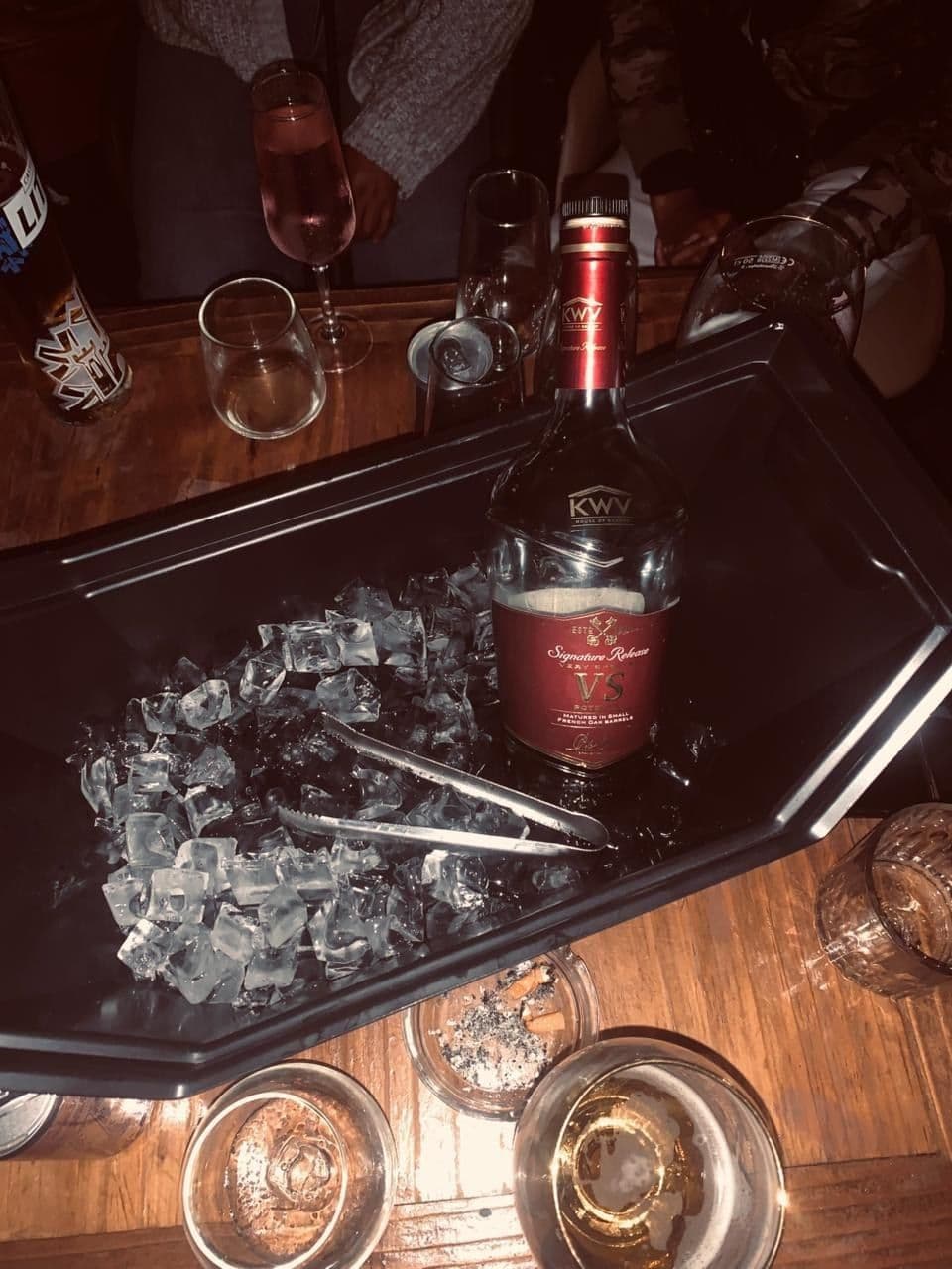 The bottle standing in a tray of ice on a dark table, tongs and tasting glasses alongside