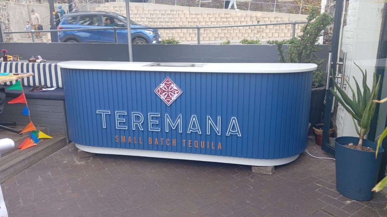A Teremana Small Batch Tequila bar unit standing outdoors on paving: a long blue slatted counter with a white top, the red diamond mark above the Teremana wordmark on its front, with planters and a striped banquette either side